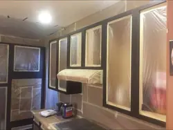 Kitchen cabinet frames finished in dark paint while the cabinet openings, range hood and surrounding wall stay masked behind plastic sheeting and taped paper.