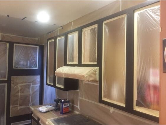 Kitchen cabinet frames finished in dark paint while the cabinet openings, range hood and surrounding wall stay masked behind plastic sheeting and taped paper.