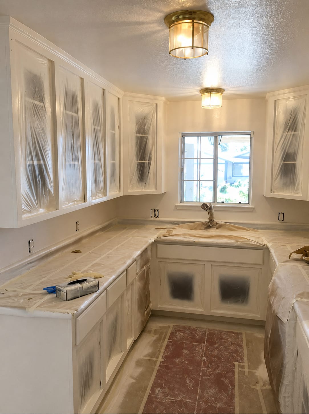 A kitchen masked for cabinet spraying. Every upper and lower cabinet panel is sheeted in plastic and taped at the edges, the countertops are covered in paper, the tap is bagged, and the tiled floor is protected.