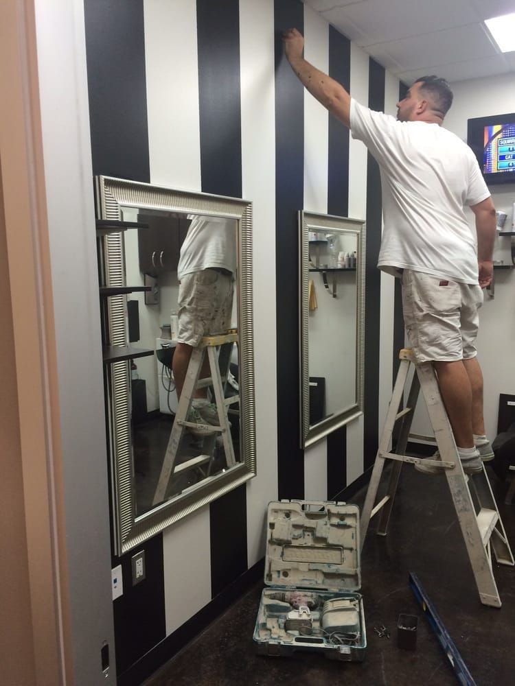 A painter on a step ladder finishing a black and white striped accent wall in a commercial salon interior, with framed mirrors mounted over the stripes and tools laid out on the floor.