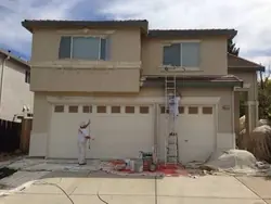Two painters working on the front elevation of a two-storey stucco home, one rolling beside the garage and one on a ladder at the upper trim, with drop cloths across the driveway.
