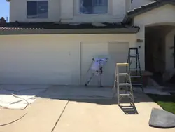 A painter spraying a garage door while the neighbouring door and the eaves above stay masked, with drop cloths laid across the driveway and a ladder to one side.