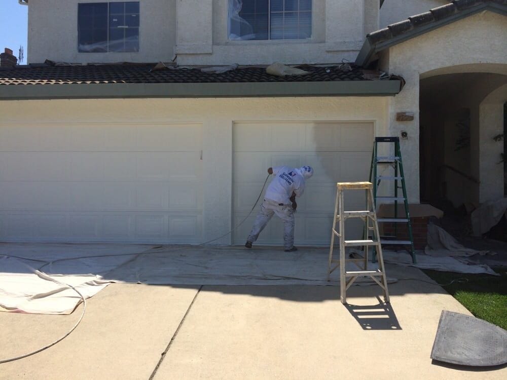 A painter spraying a garage door while the neighbouring door and the eaves above stay masked, with drop cloths laid across the driveway and a ladder to one side.