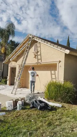 A painter spraying the gable and fascia of a stucco home from the driveway, with the roof edge masked in paper and drop cloths laid across the path and entry.