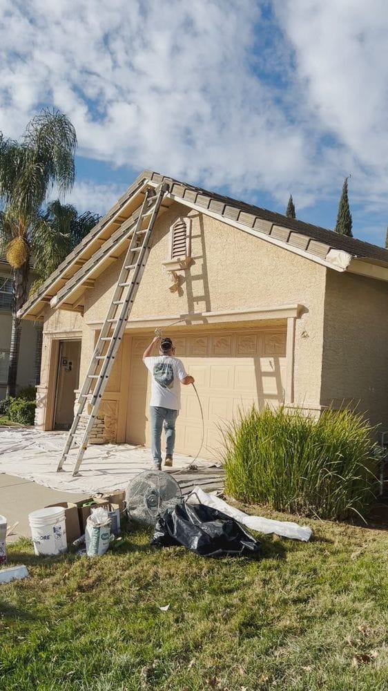 A painter spraying the gable and fascia of a stucco home from the driveway, with the roof edge masked in paper and drop cloths laid across the path and entry.