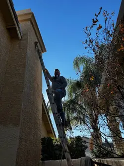 A painter in a respirator working from an extension ladder at the eaves of a stucco house, cutting in the fascia against a clear sky.