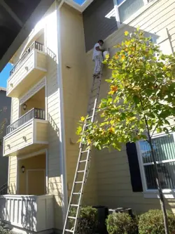 A painter working from a tall extension ladder on the upper siding and trim of a three-storey building with balconies.