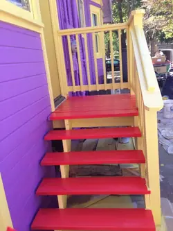 Finished exterior stairs and porch of a Victorian house, with red-painted treads and landing, pale yellow railings, posts and trim, and purple siding.