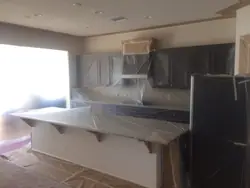A Folsom kitchen masked before spraying. Plastic is taped over every cabinet door and the range hood, the island and countertops are sheeted, and paper covers the floor, leaving only the walls and ceiling exposed.