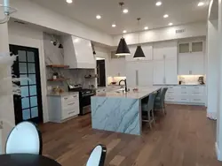 A finished open-plan Folsom kitchen with white cabinetry to the ceiling, a marble-topped island, a shaped range hood and dark pendant lights over the counter.