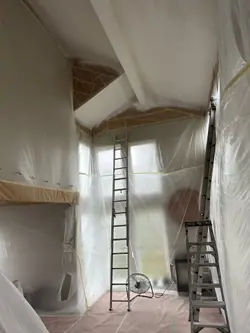 A double-height room masked from floor to ceiling before painting. Plastic sheeting covers the walls and windows, paper is taped along the ceiling line and around the beams, the floor is covered, and an extension ladder is set for the vaulted section.