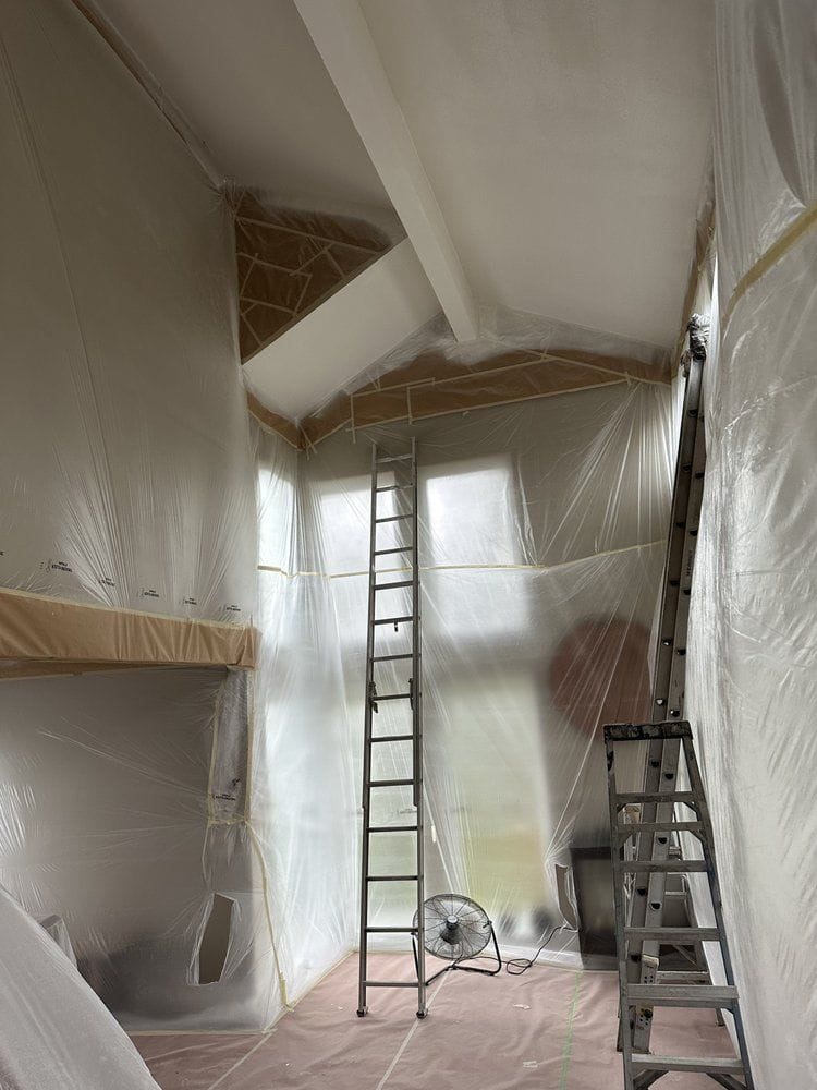 A double-height room masked from floor to ceiling before painting. Plastic sheeting covers the walls and windows, paper is taped along the ceiling line and around the beams, the floor is covered, and an extension ladder is set for the vaulted section.