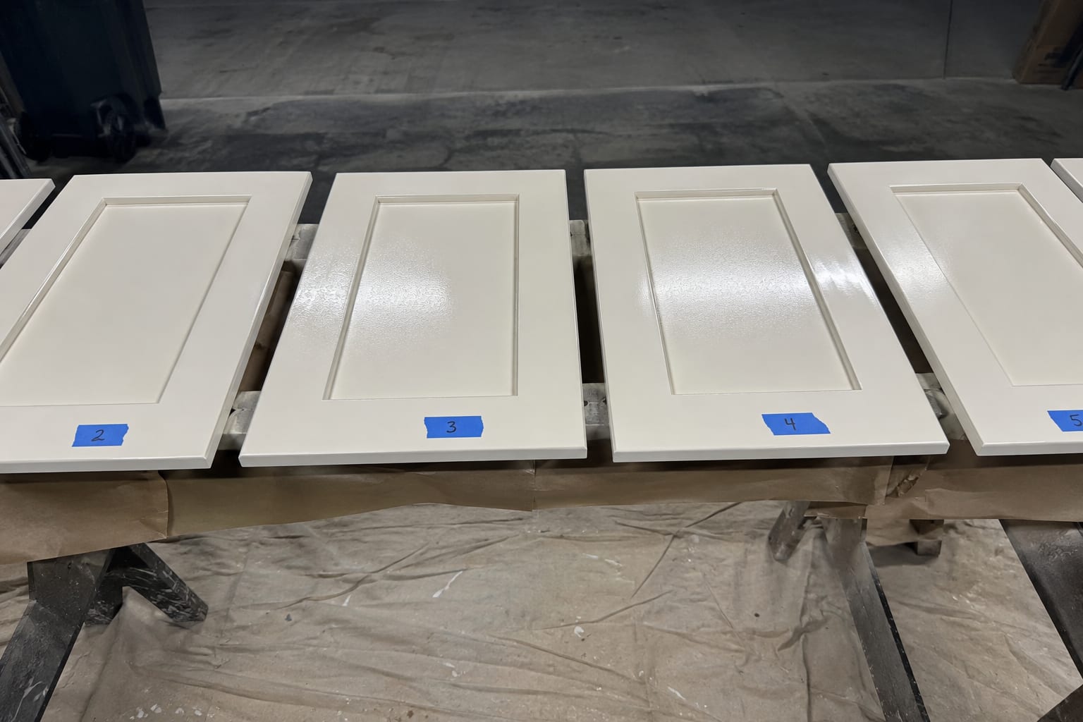 Freshly sprayed cabinet doors laid out flat on rails above a sheeted floor, each one numbered with a strip of blue tape so it goes back where it came from.