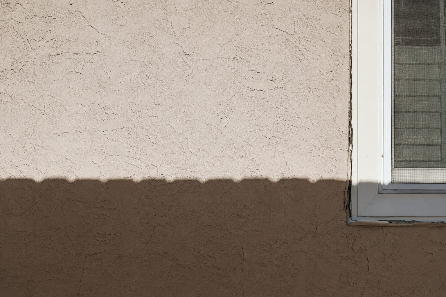 A stucco wall in hard afternoon sun, the lit upper section pale and the shaded lower section much darker, with hairline cracks across the render and split caulking running down the edge of the window frame.
