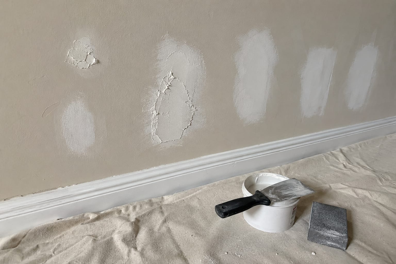 A wall part-way through preparation, with several patches of white filler spread over dents and nail holes, and a filling knife, tub and sanding block on the drop cloth below.