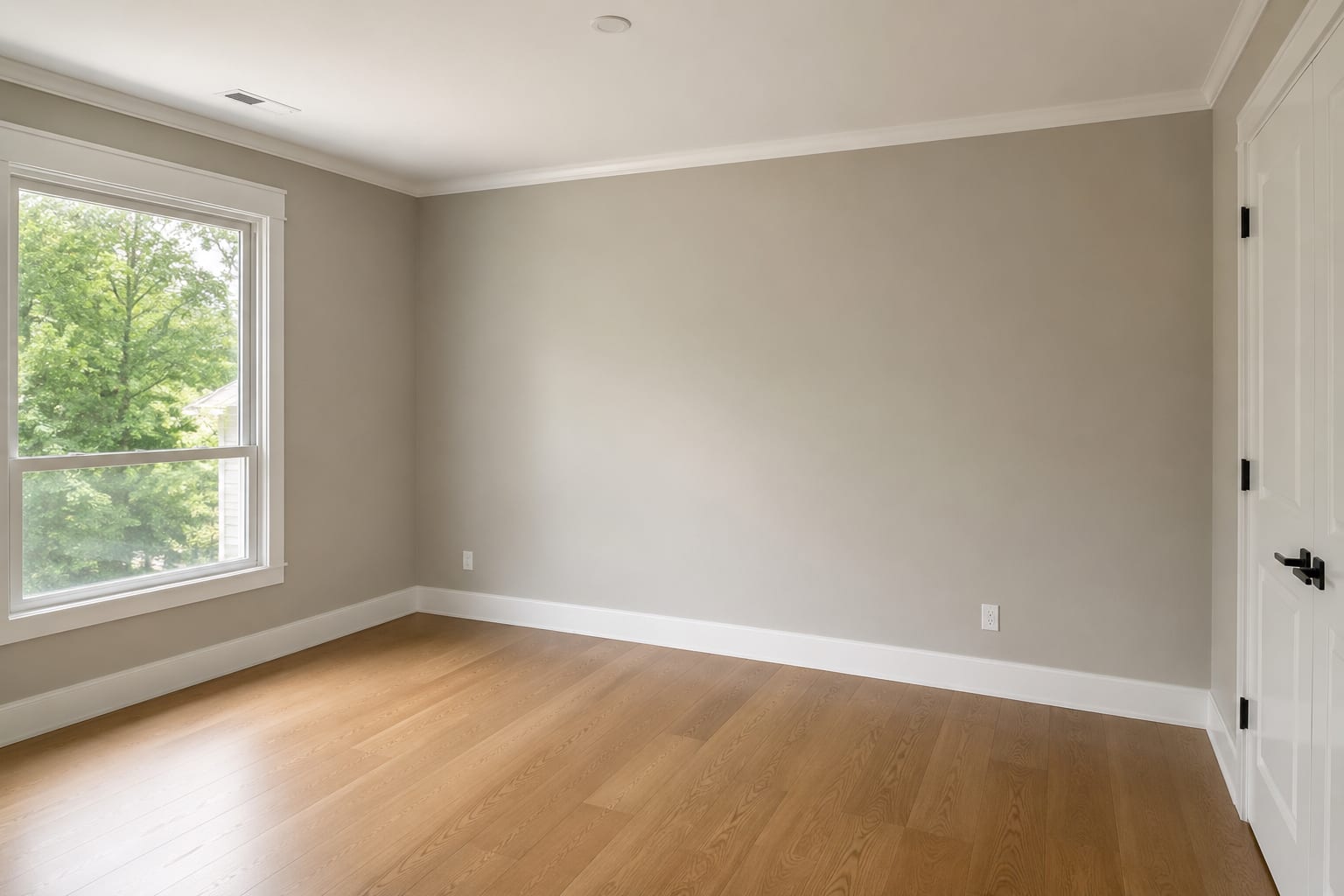 An empty room after painting, with plain grey-beige walls, white skirting, door casing and coving, and a wood floor lit by a single window.