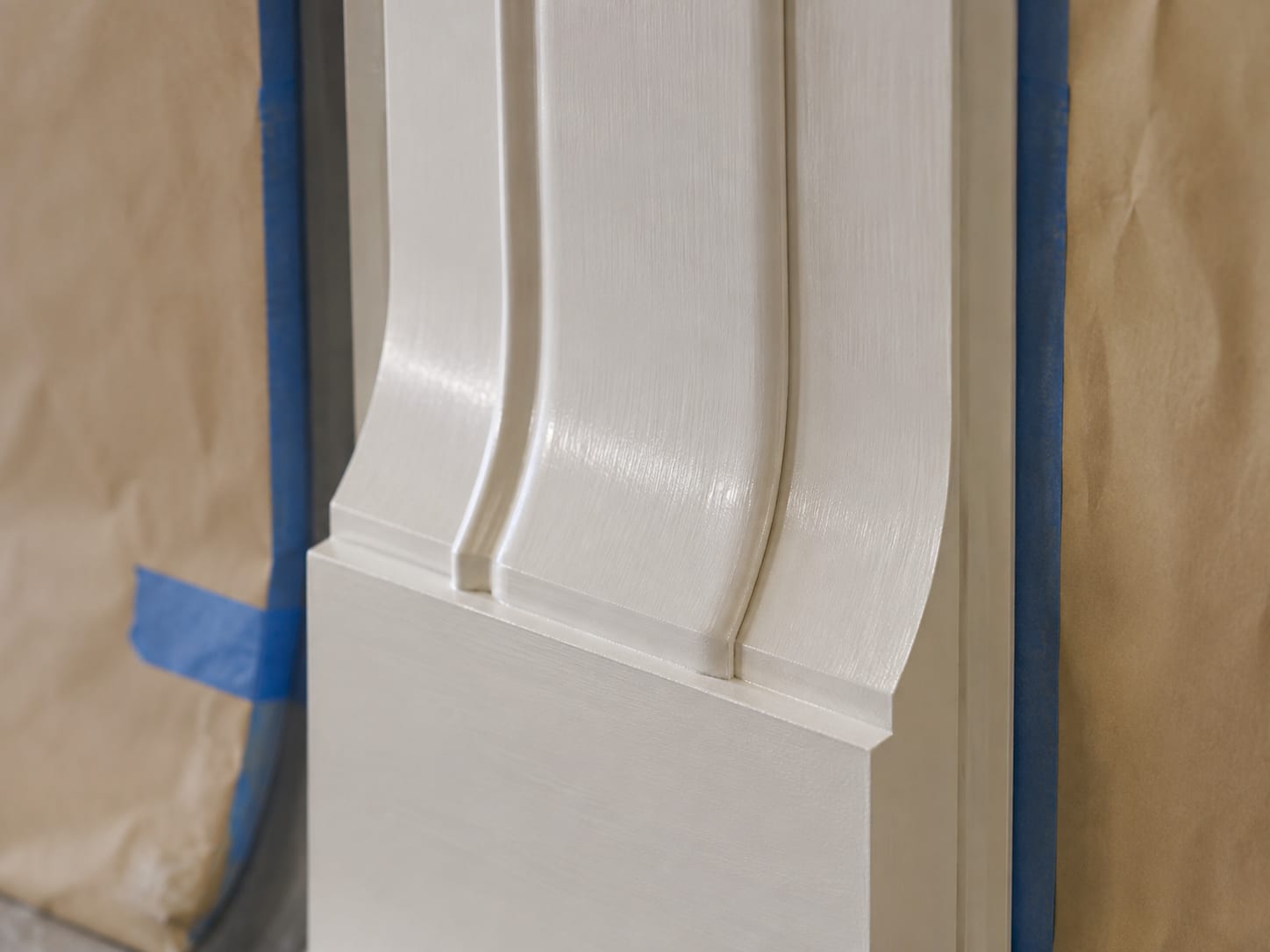 A close-up of painted trim between two masked areas, with blue tape and brown paper protecting the surfaces on either side of a crisp painted edge.