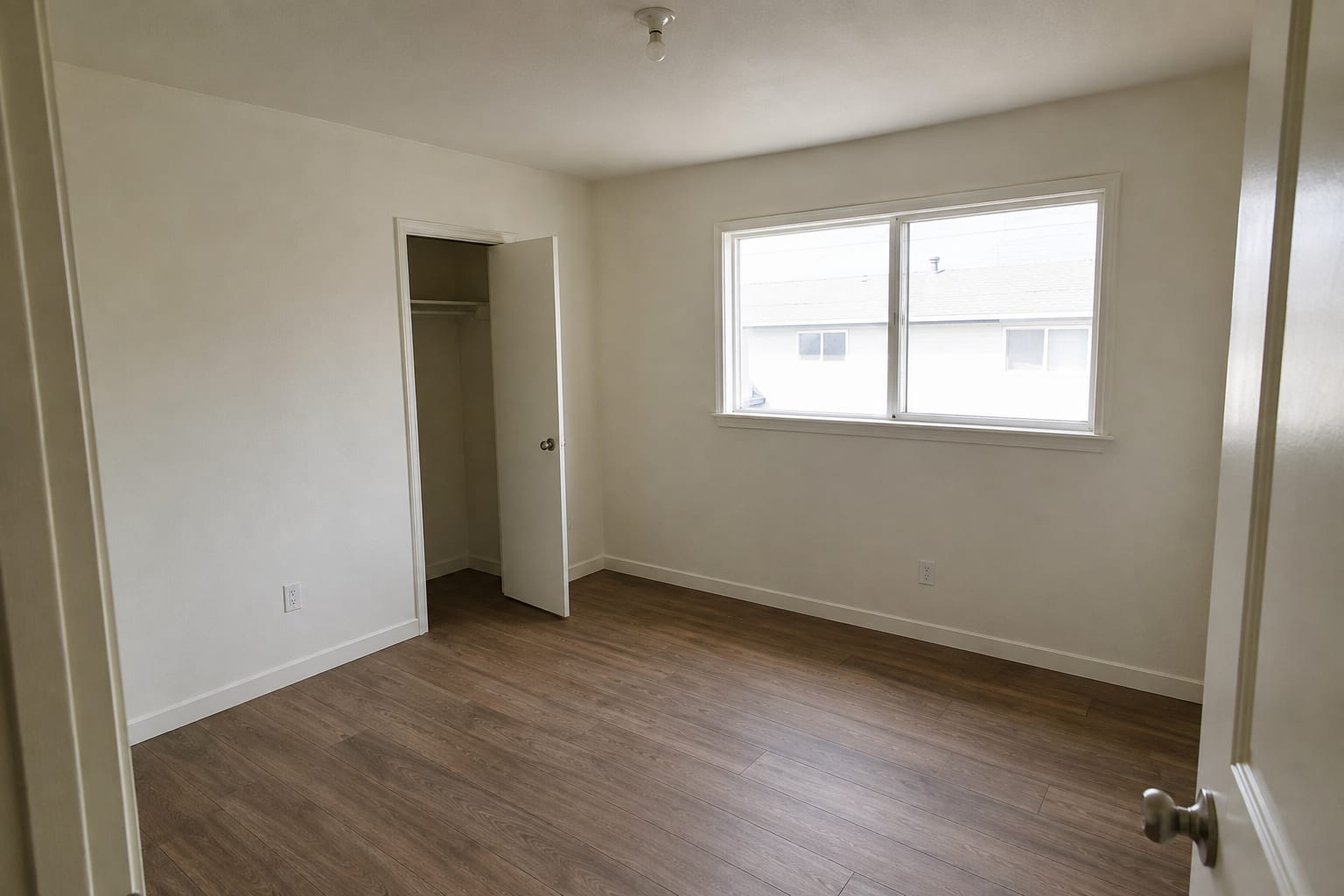 An empty rental room repainted in plain off-white, with white trim, a wardrobe standing open, a bare ceiling fitting and new wood-effect flooring.