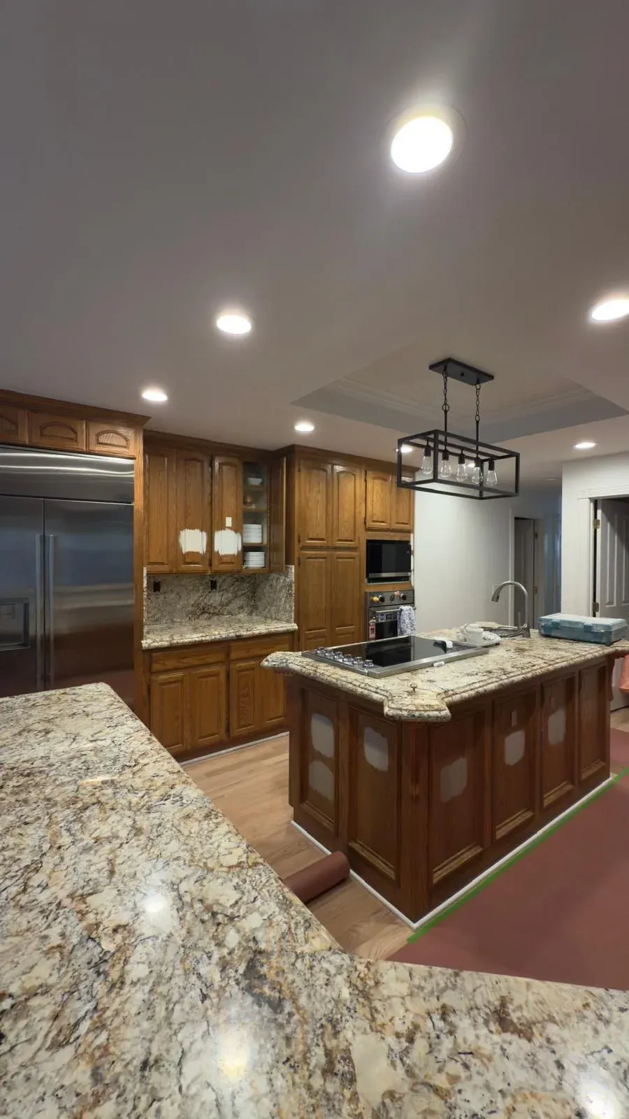 A wide-angle interior view of a renovated kitchen with two large granite-topped islands and a distinctive dark metal pendant chandelier.